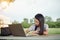 Young female university student writes a note and assignment with a laptop beside outside the campus with blur green tree backgrou