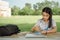 Young female university student writes a note and assignment with a laptop beside outside the campus with blur green tree backgrou