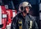 Young fireman wearing protective uniform standing next to a fire engine in a garage of a fire department