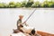 Young fisherman in camo looking at thirsty dog, while angling in calm river from boat.