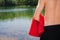 Young and fit, athletic male man standing on the lake shore without a shirt while holding red towel. Swimming, summer activities
