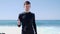 Young fit attractive handsome man in black showing thumbs up to the camera standing on the beach. Strong waves are hitting the bea