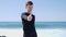 Young fit attractive handsome man in black showing thumbs up to the camera standing on the beach. Strong waves are hitting the bea