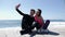 Young fit couple sitting near the beach holding phone and taking selfies posing. Happy boyfriend and girlfriend taking pictures. S
