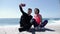 Young fit couple sitting near the beach holding phone and taking selfies posing. Happy boyfriend and girlfriend taking pictures. S