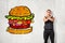 Young fit man in sleeveless hoody standing with arms crossed across chest at white wall with drawing of huge hamburger