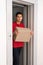 Young friendly delivery courier dressed in red polo shirt leaving the elevator holding cardboard box with order