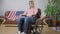 Young frustrated American woman sitting in wheelchair in living room at home with national flag on couch at background