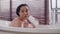 Young funny pretty hispanic woman sitting in bath bathing singing song using shampoo bottle as microphone happy