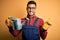 Young gardener man wearing working apron and gloves holding watering can very happy pointing with hand and finger to the side