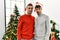 Young gay couple standing by christmas tree wearing hat winking looking at the camera with sexy expression, cheerful and happy