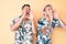 Young gay couple of two men wearing summer hat and hawaiian shirt shouting angry out loud with hands over mouth