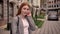 Young ginger female student standing in city street, looking in camera and touching her hair, smiling, building