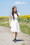 A young girl of 17-20 years old is walking in a white dress along the road along a field with a blooming sunflower. Concept: a rur