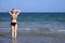 Young girl in a bathing suit and a striped hat is standing in the sea holding her hands behind her head, fingers folded into the l