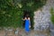 A young girl in a beautiful long blue dress stands near the old castle, the old ruins near the green dense bushes stands with her