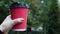 A young girl in a coat holds morning coffee with her while walking in the park. Hand holding paper cup of coffee in green park.