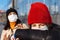 Young girl coughs in an elbow to cover. Two girls waiting for a bus. Woman leans back from a sneezeing girl on a tram station.