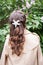 Young girl with curly hair is standing near blooming lilac bush and looking at it. Rear View.