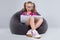 Young girl in glasses doing homework on a gray bean bag