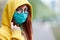 A young girl with glasses, a reusable medical mask, and a yellow hood turns and looks around