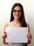 A young girl in glasses shows a blank sheet. Colorful portrait on a white background. Space for text. The brunette is smiling