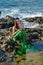 A young girl in a green mermaid costume with red hair sits on the rocks in the water and looks at the shore.