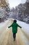 A young girl in a green sweater and hat walks in the middle of a snowy road in a thick pine forest. Freezing day