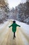 A young girl in a green sweater and hat walks in the middle of a snowy road in a thick pine forest. Freezing day