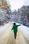 A young girl in a green sweater and hat walks in the middle of a snowy road in a thick pine forest. Freezing day.