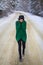 A young girl in a green sweater stands in the middle of a snowy road in a thick pine forest. Freezing day