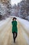 A young girl in a green sweater stands in the middle of a snowy road in a thick pine forest. Freezing day.