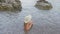 Young girl in hat ans swimsuit sitting in sea water on pebble beach at summer holiday. Tourist girl enjoying soft waves
