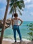 A young girl in jeans and a white T-shirt stands and looks at the sea under a palm tree