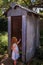 A young girl with long red hair and a blue dress curiously opens the door of an rustic outdoor toilet typically used in Australia