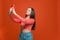 Young girl making selfie on phone. Pretty woman making faces on camera, posing for picture on orange studio background