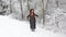 A young girl with red long hair in a black dress and hat stretches up while standing in a snow covered forest