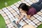 A young girl in a red T-shirt and straw hat lies on a plaid, on green grass, and works behind a laptop remotely. Telework