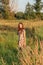 A young girl in a retro vintage dress with flowing hair is standing in a tall grass in a sunlight afterglow with a suitcase