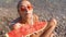 Young girl sits on sea beach, bites off large piece of watermelon