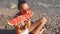 Young girl sits on sea beach, bites off large piece of watermelon