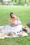 Young girl sitting on plaid near fruits and hat, eating watermelon, grass in background.