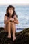 Young girl sitting on the rock near the ocean and smiling. Happy childhood. Spending time on the beach. Cute little girl. Summer