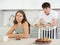 Young girl sitting at the table while her boyfriend is quarreling with her standing behind