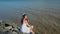 Young girl in straw hat sitting on stone beach. he closes his eyes, enjoys sun.