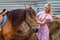 Young girl stroking a head of brown horse before a walk that eats hay near the haystack on a summer clear day