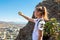 Young girl on top of rock takes selfie using an action camera against background of landscape with blue sky, city and mountains