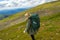 Young girl travel with big green backpack go down the hill on the rocks towards the green fields in altai mountains named sarlyk,