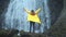 Young girl traveler hiker in a yellow raincoat walks to a waterfall in the highlands, raises his hands up, enjoys nature