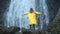 Young girl traveler hiker in a yellow raincoat walks to a waterfall in the highlands, raises his hands up, enjoys nature